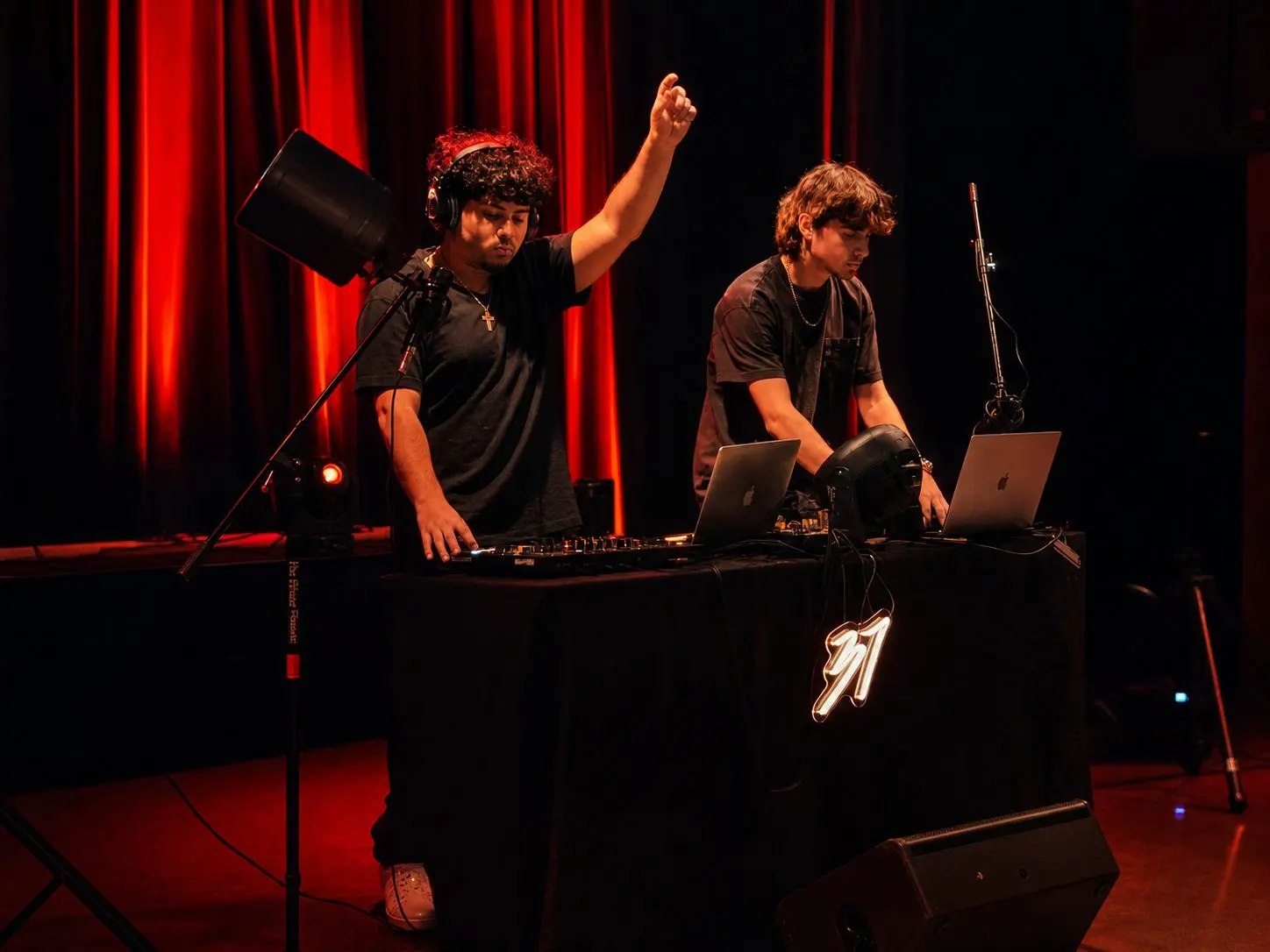 Two DJs behind the DJ Studio 37 booth on a stage, a red curtain lit behind them