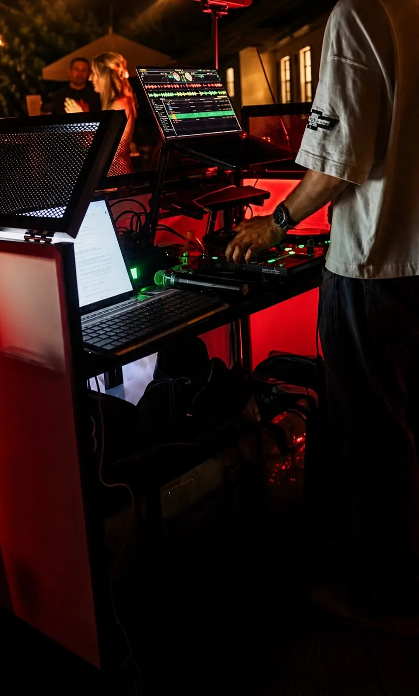 A DJ’s hands on the controller at night, a waveform on one screen and the running order on another